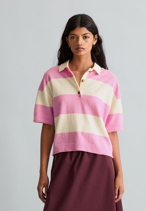 Junge Frau mit langem dunkelhaar trägt ein rosa-creme gestreiftes Polohemd mit kurzen Ärmeln und einen dunkelburgunderfarbenen Rock vor einem grauen Hintergrund.