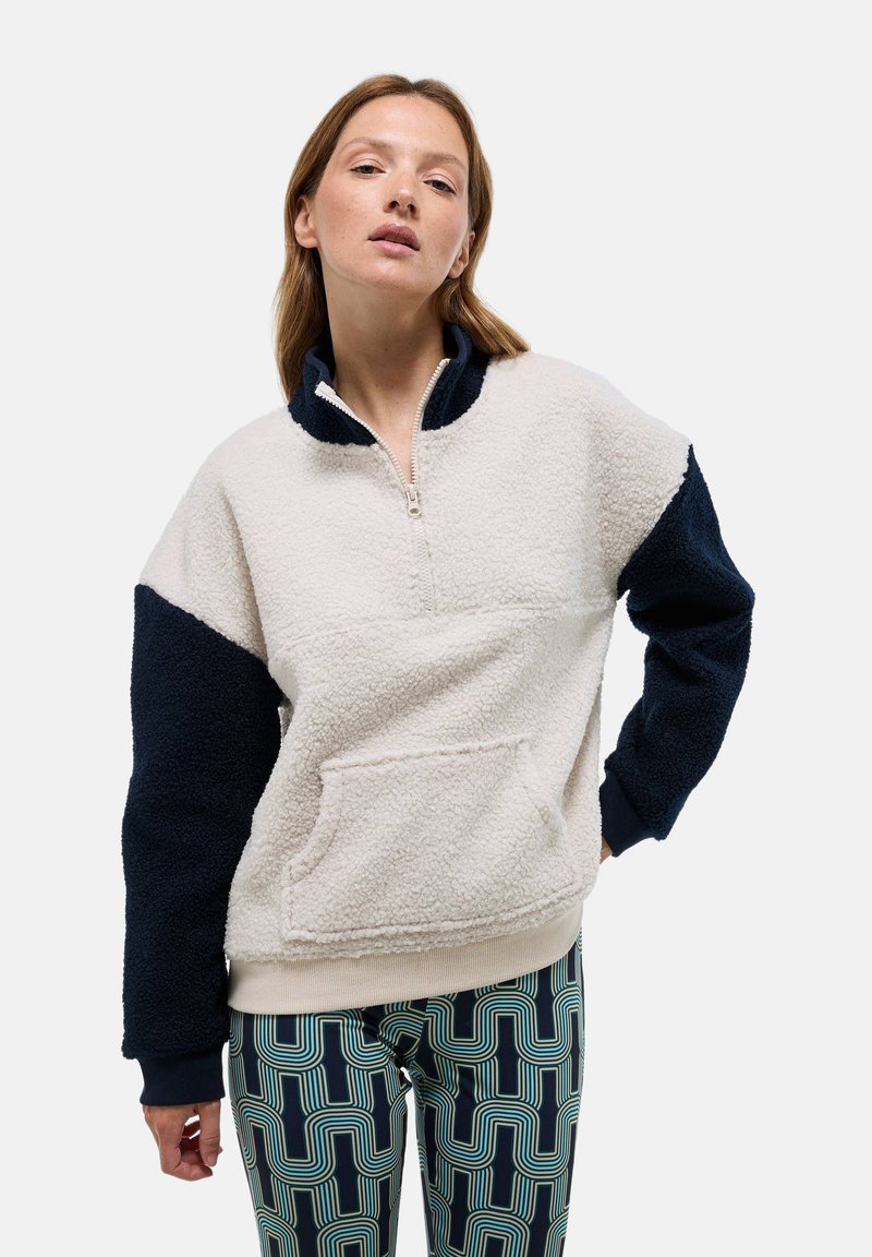 Pull en polaire avec demi-zip, corps crème, manches bleu marine, poche avant, tissu texturé et poignets côtelés. Associé à des leggings à motifs.