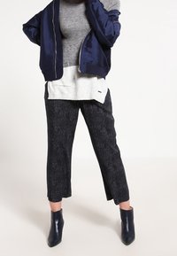 Marineblauer Bomberjacke, grauer Pullover darunter, graue und schwarz gemusterte cropped Hose und dunkelblaue Stiefeletten. Glatte und strukturierte Stoffe.