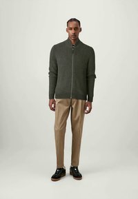 Man med en mörkgrön zip-up-ribbad tröja, beige byxor och svarta sneakers med vita sulor, stående mot en enfärgad ljus bakgrund.