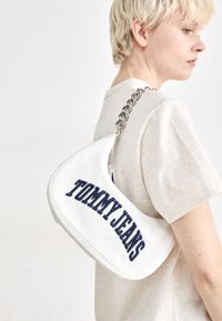 Balta pečių rankinė su kreivumu, dekoruota mėlyna išsiuvinėta "TOMMY JEAN'S" užrašu ir sidabriniu grandininiu dirželiu. Lygi tekstūra.