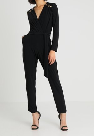 Vrouw die een zwarte jumpsuit met lange mouwen draagt, met gouden knopen op de schouders, een wikkelriem en zwarte open teen hoge hakken.