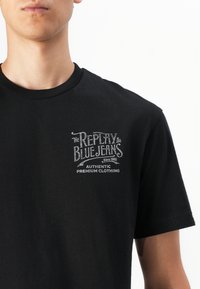 Black cotton t-shirt with a round neck, featuring a silver graphic print that includes the brand name "Replay" and "Blue Jeans" text.