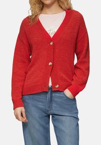 Rode gebreide cardigan met V-hals, lange mouwen en vijf knopen. Heeft geribde manchetten en onderkant. Draagt over een licht shirt, gecombineerd met blauwe jeans.