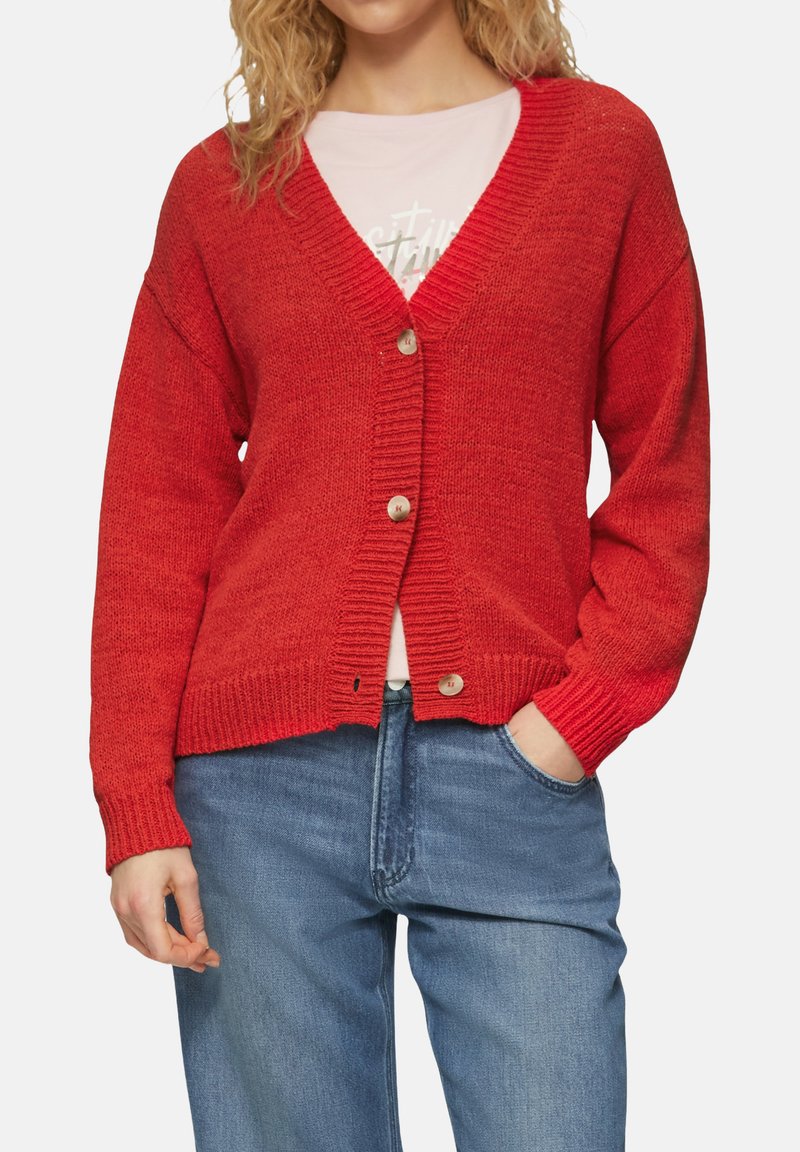 Rode gebreide cardigan met V-hals, lange mouwen en vijf knopen. Heeft geribde manchetten en onderkant. Draagt over een licht shirt, gecombineerd met blauwe jeans.