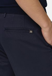 Nahaufnahme einer Person, die marineblaue Hose mit einer hinteren Leistentasche und einem kleinen schwarzen Etikett mit weißem Buchstaben "B" trägt.