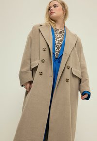 Abrigo de lana beige con solapas oversize, doble botonadura y bolsillos frontales, superpuesto sobre una prenda azul y una blusa estampada.