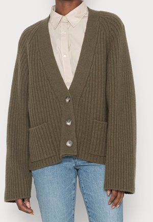 Personne portant un cardigan côtelé vert olive surdimensionné avec poches avant et boutons, par-dessus une chemise beige à col et un jean bleu clair.