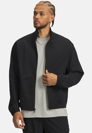 Must tikk-kinnitatud jakk tekstuurse ülaosaga ja võrkriidest paneelidega, millel on elastsed varrukasäärised ja kerge läige. Kantud üle halli T-särgi.