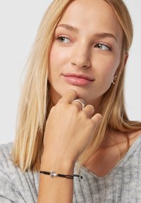 Pulsera negra ajustable con un charm de estrella de plata y un acento de perla, llevada en una mano, combinada con un anillo de plata, sobre un suéter gris texturizado.