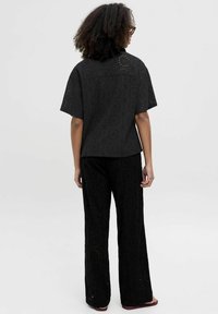 Persona vistiendo una blusa de manga corta con patrón de ojales negros y pantalones de pierna ancha a juego, de pie con la espalda hacia la cámara sobre un fondo liso.