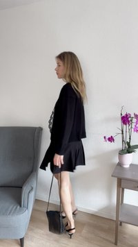 Outfit nero con un top strutturato e una gonna svasata. Accompagnato da una piccola borsa nera e sandali con tacco. Pavimento in legno chiaro e sedia grigia.