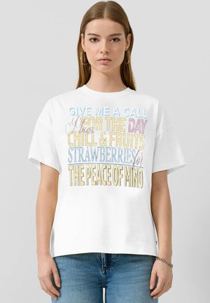 Młoda kobieta o prostych włosach nosi białą koszulkę z kolorowym napisem o relaksie, owocach, truskawkach i spokoju ducha, w połączeniu z niebieskimi dżinsami.