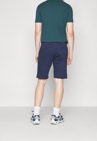 Tmavě modré šortky vyrobené z bavlny, s klasickým střihem a ohrnutými lemy. Spárované s zelenou košilí s krátkým rukávem a sportovními botami.