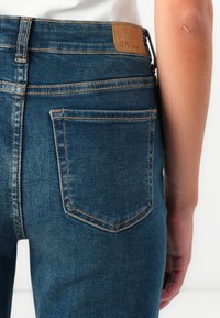 Mørkeblå denimjeans med teksturert stoff, med et brunt skinnmerke, baklomme og kontrastfarget oransje søm.