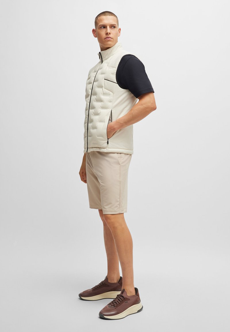Gilet en crème rembourré avec fermeture éclair à l'avant et accents noirs, associé à un short beige clair et des baskets marron. Texture lisse, design décontracté.