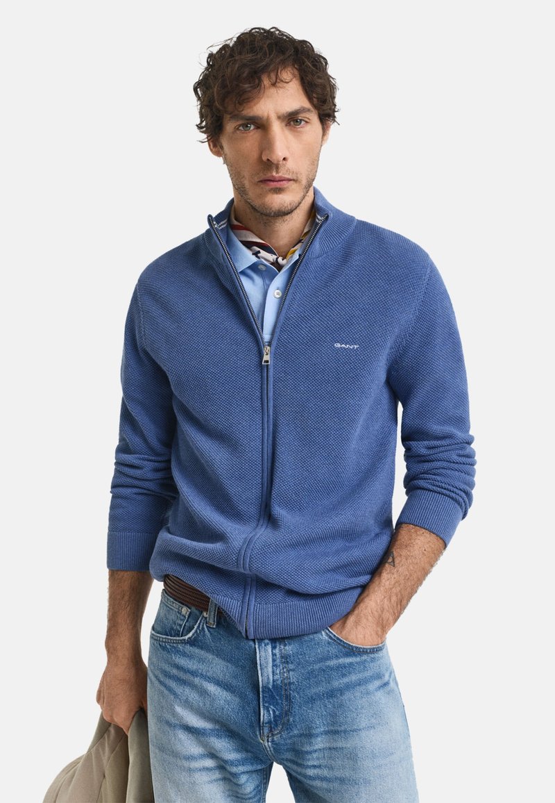 Blauer, gestrickter Reißverschluss-Pullover mit strukturiertem Muster, gerippten Bündchen und Kragen. Getragen über einem hellblauen Poloshirt und kombiniert mit Jeans.