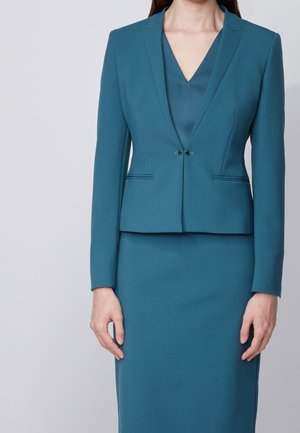 Kvinde iført en tætsiddende teal blazer med dobbelthætteknapper foran og matchende teal pencilskørt, stående mod en ensfarvet lys baggrund.