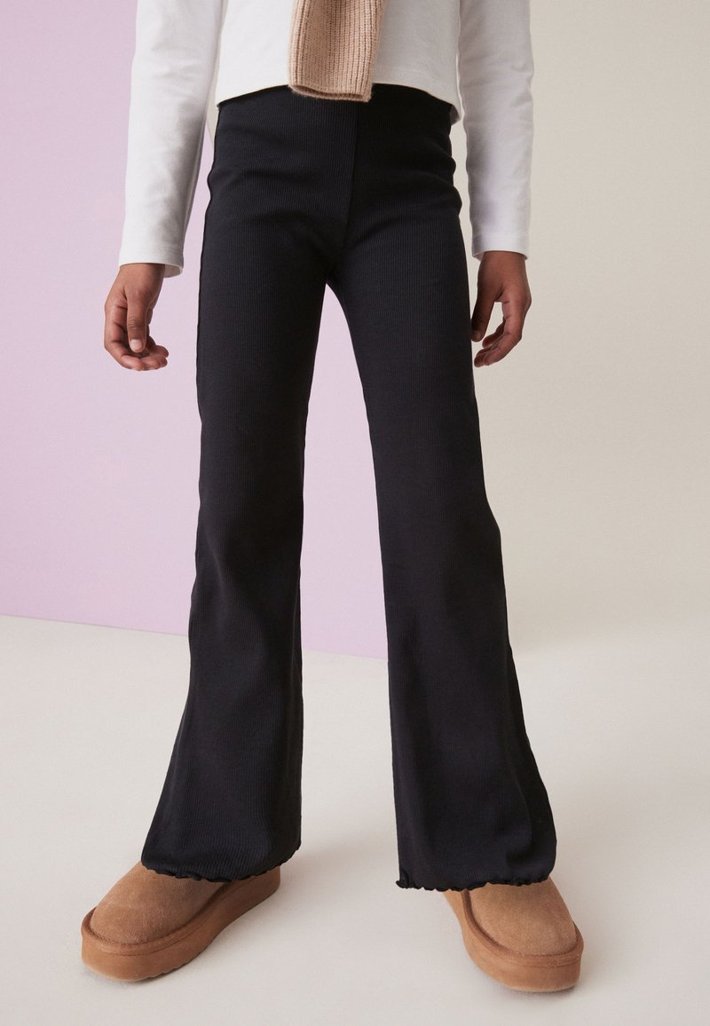 Persona con pantalones negros acampanados, camiseta blanca de manga larga, bufanda beige y zapatos tipo slip-on marrones, de pie sobre un suelo claro.