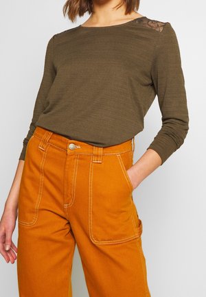 Kobieta ubrana w oliwkowo-zieloną bluzkę z długim rękawem i koronkowym detalem na ramionach oraz jasnopomarańczowe, wysokowarstwowe spodnie z dużymi przednimi kieszeniami.