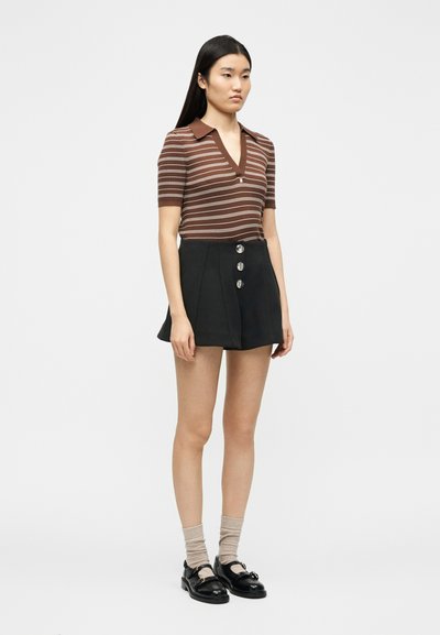 Γυναίκα με μακριά σκούρα μαλλιά που φοράει καφέ ριγέ τοπ με κοντά μανίκια, μαύρο ψηλόμεσο σορτς με κουμπιά, μπεζ κάλτσε ς και μαύρα παπούτσια.