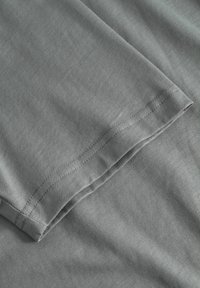 Szürke pólóanyag, sima textúrával és egy hajtott ujjrésszel, rajta egyenes varratdíszítéssel az alsó szegély mentén.