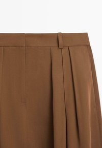 Brązowe spodnie z wysokim stanem, z zakładkami, szlufką na pasek oraz widocznym przeszyciem wzdłuż pasa i przodu.