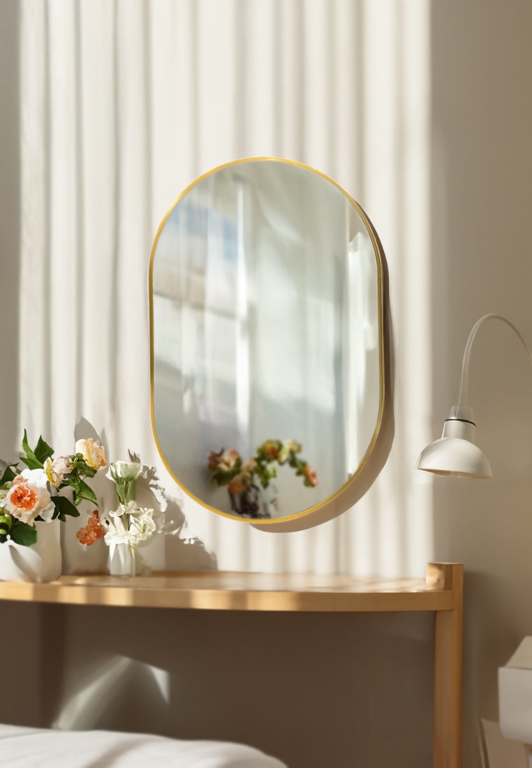 Ovaler Spiegel mit einem dünnen, goldfarbenen Rahmen. Glatte, reflektierende Oberfläche umgeben von einem sanft geschwungenen Kontur. Minimalistisches Design.