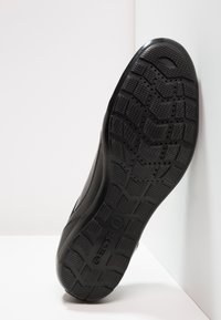 Adidași negri cu o talpă din cauciuc texturat, având un model hexagonal. Partea superioară este netedă, concepută pentru uz casual.