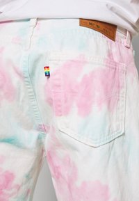 Jeans blancs avec un motif tie-dye rose et bleu clair, dotés d'une poche arrière et d'une petite étiquette logo multicolore près de la poche.