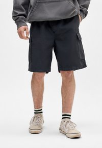 Shorts cargo noirs avec poches latérales, en coton et à coupe décontractée. Portés avec des baskets blanches et des chaussettes rayées vertes.