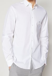 Hombre vestido con una camisa blanca ajustada de botones, con un pequeño logo negro "ck" en el pecho, combinado con pantalones gris claro y manos en los bolsillos.