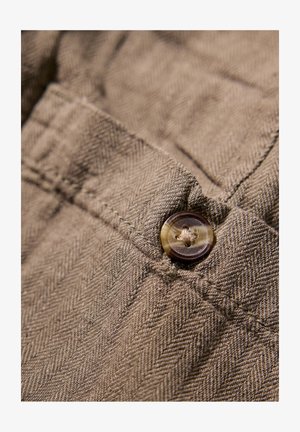 Primer plano de una tela marrón de espiga con un botón redondo oscuro y detalles de costura visibles. La textura parece suave y tejida.