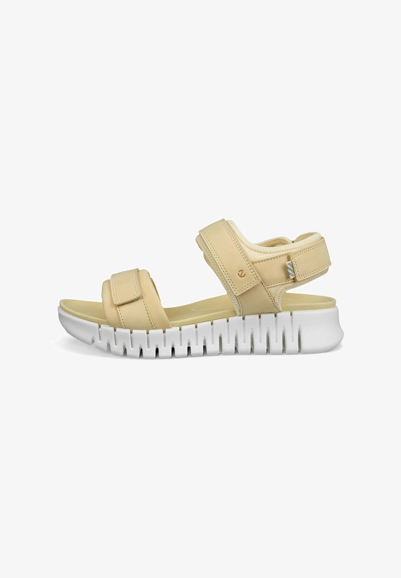 Beige sandal with adjustable straps and white elevated sole featuring a scalloped pattern for traction. Soft material and cushioned footbed.