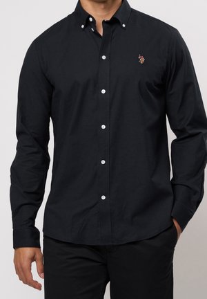 Homme portant une chemise noire à manches longues avec un petit logo brodé de joueur de polo sur la poitrine, pantalon noir, une main dans la poche.