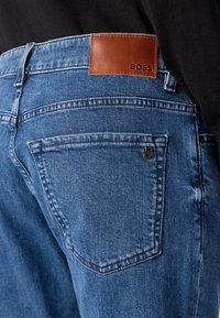 Mėlyni džinsai iš džinsinio audinio su odiniu lipduku su užrašu „BOSS“ nugaroje. Yra standartinės kišenės ir siūlių detalės.