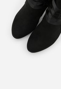 Botas negras hasta el tobillo de ante con una textura suave, punta afilada y acentos de cinta alrededor del tobillo.