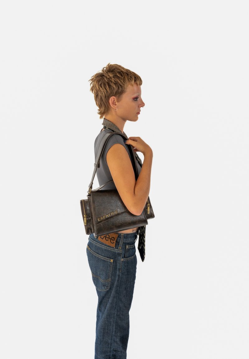 Barna bőr kézitáska cipzár díszítéssel, téglalap alakú, és az "EXPATRIÉ" logóval. A modellen szürke felső és farmernadrág van.