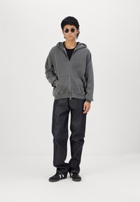 Grå zip-up hoodie med kängurufickor, kombinerad med mörka jeans och svarta sneakers med vita ränder. Modellen bär solglasögon.