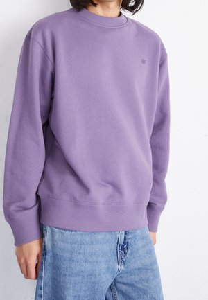 Person trägt ein schlichtes violettes Sweatshirt mit Rundhalsausschnitt und hellblaue Denimjeans, gezeigt von der Mitte der Brust bis zu den Hüften vor weißem Hintergrund.