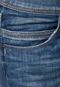 Jeans de mezclilla azul con un bolsillo frontal que muestra múltiples líneas de costura y un pequeño botón metálico a un lado. La textura parece ligeramente desgastada.