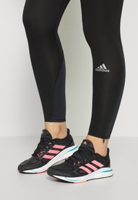 Zwarte sportlegging met een rits bij de enkel, gecombineerd met zwarte hardloopschoenen met roze strepen en witte zolen.