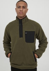 Olive green fleece pullover with a black zippered chest pocket, side pockets, high collar, and textured fabric. Features raglan sleeves.