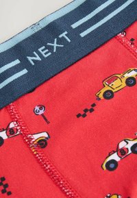 Röda bomullsunderkläder med ett blått elastiskt midjeband som har "NEXT"-logotypen, dekorerade med små färgglada bil- och stoppskyltmönster.