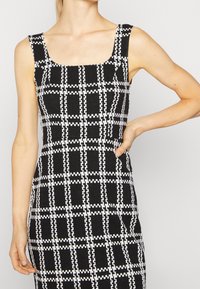 Schwarz-weiß kariertes Kleid mit quadratischem Ausschnitt, ärmellosem Design und taillierter Form; strukturiertem Stoff mit einem gitterartigen Muster.