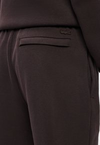 Tmavohnedé nohavice s textúrovanou tkaninou, úzkym strihom a jedným zadným vreckom s nenápadným logom v rovnakej farbe.