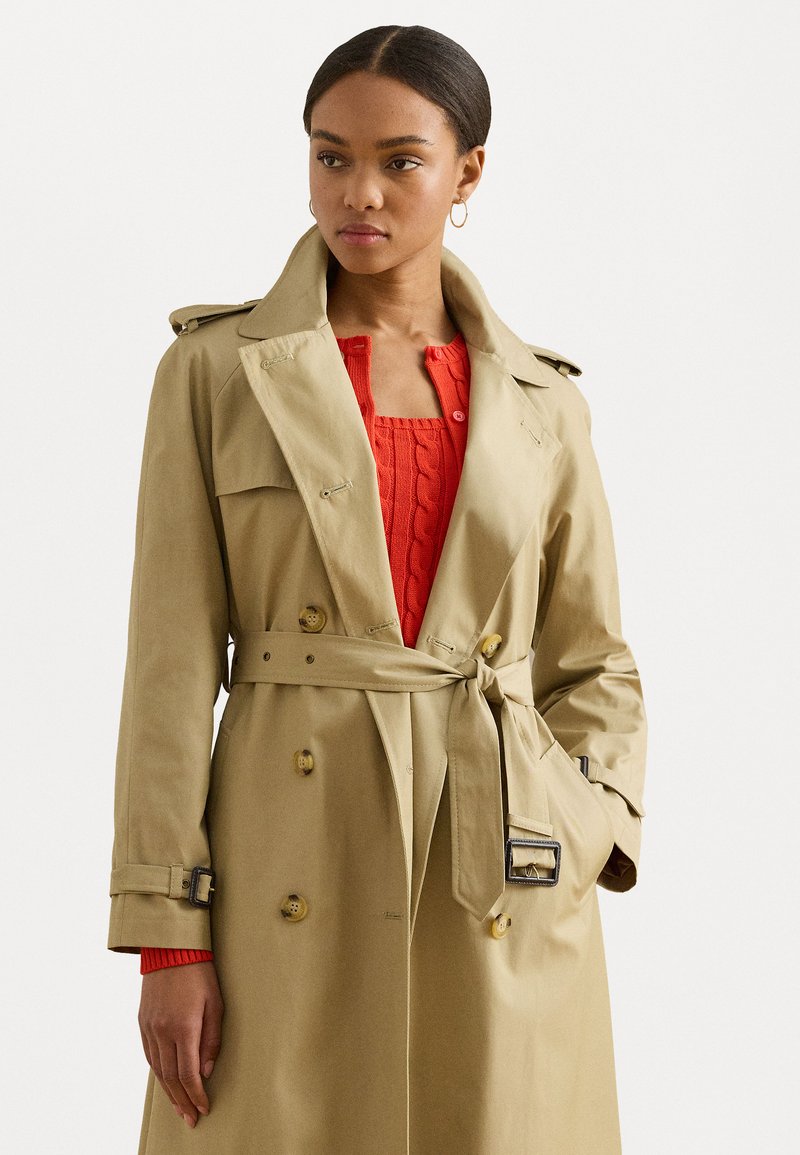 Beige kaksirivinen trenchcoat, jossa on vyö, olkapään hihnat ja nappi yksityiskohtia. Päällä punainen nyöri-neulepaita.