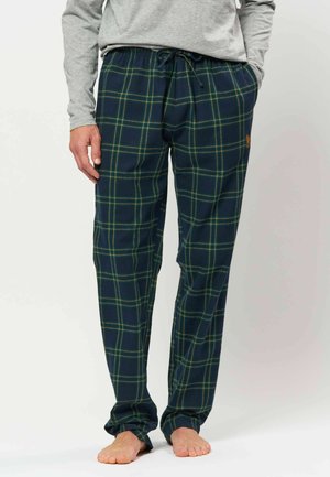 Homme portant un pantalon de pyjama à carreaux bleu et vert avec une chemise grise à manches longues, debout pieds nus les mains dans les poches.