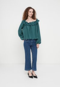 Τιρκουάζ μπλούζα έξω από τον ώμο με βολάν στον λαιμό, μακριά μανίκια και άνετη γραμμή, σε συνδυασμό με κοντά μπλε τζιν και μαύρα παπούτσια με λουράκι στο πίσω μέρος.