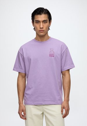 Un homme porte un T-shirt lavande orné d’un petit motif « Desert Trek », associé à un pantalon crème, sur un fond blanc uni.